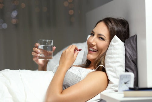 A woman in bed smiling while holding a glass of water and a pillbox, with a pillow on her head and a nightlight on in the background.